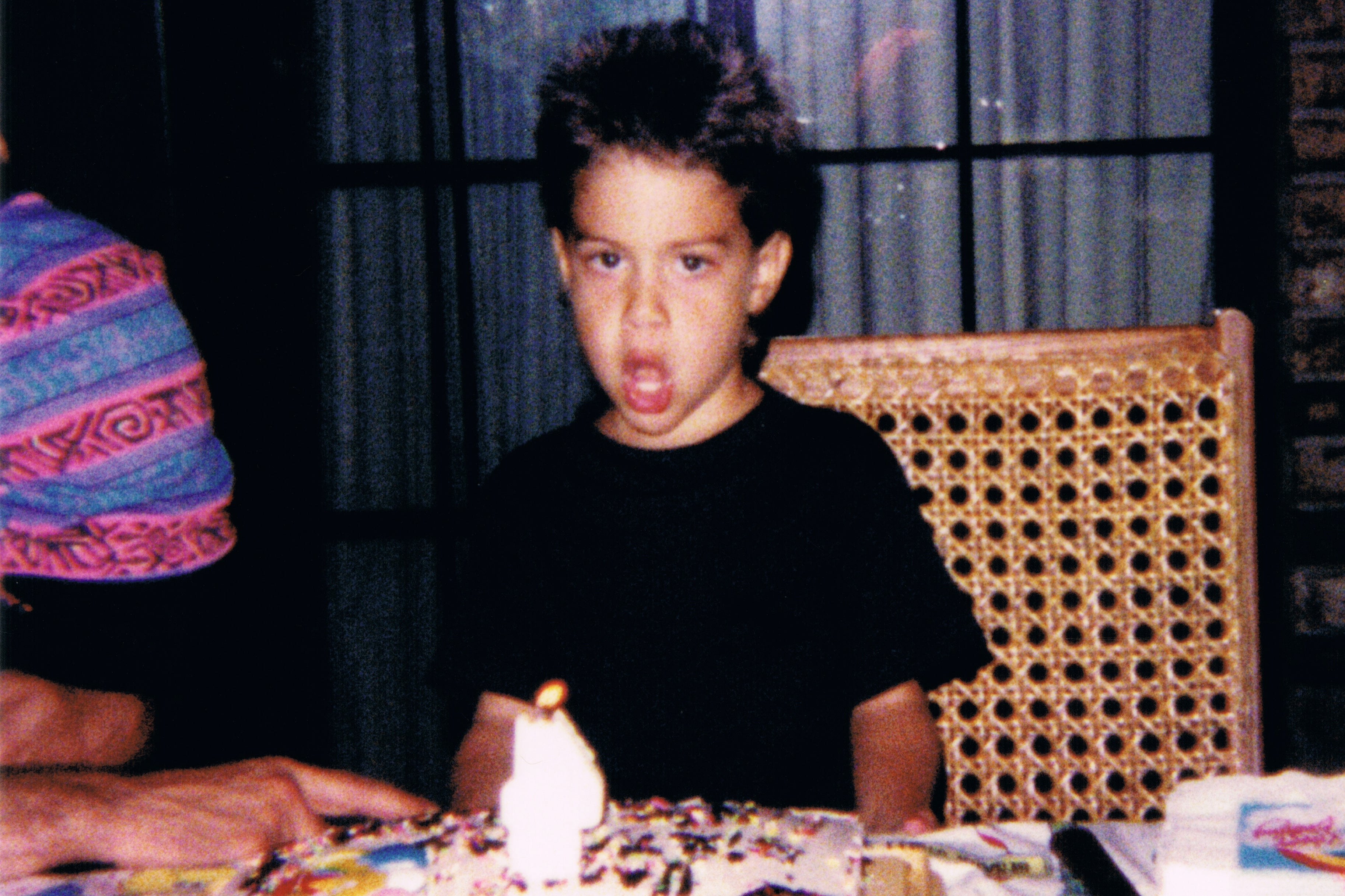 Child making a surprised expression at a birthday party with a cake and lit candle.